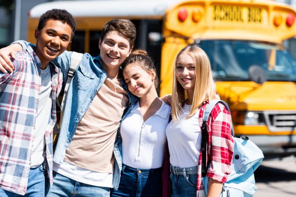 group of teen happy scholars looking at camera and embracing while standing in front of school bus